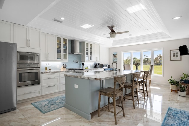 kitchen with white cabinetry, stainless steel appliances, a tray ceiling, an island with sink, and wall chimney exhaust hood