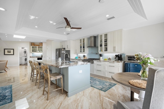 kitchen featuring a kitchen island with sink, stacked washer / dryer, light stone countertops, wooden ceiling, and wall chimney exhaust hood