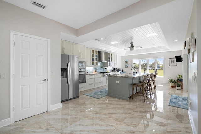 kitchen featuring a breakfast bar area, appliances with stainless steel finishes, a kitchen island with sink, light stone countertops, and wall chimney exhaust hood