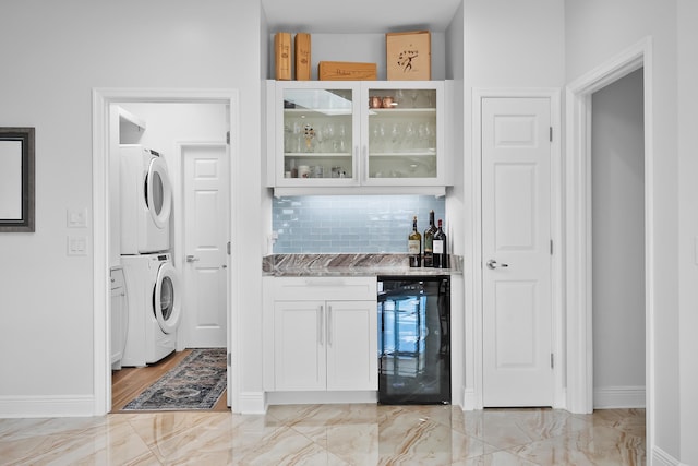 bar featuring white cabinetry, stacked washing maching and dryer, beverage cooler, light stone countertops, and backsplash