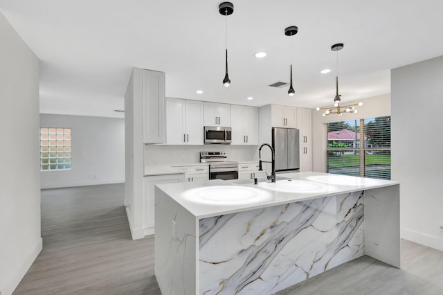 kitchen with appliances with stainless steel finishes, an island with sink, pendant lighting, light stone countertops, and white cabinets