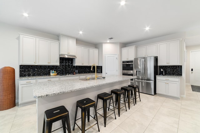 kitchen with sink, appliances with stainless steel finishes, white cabinetry, light stone countertops, and an island with sink