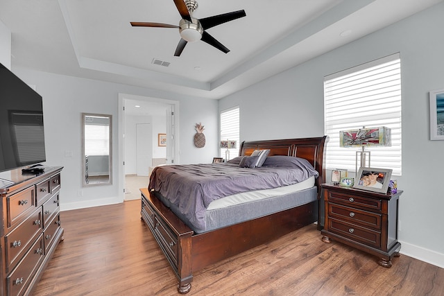 bedroom with wood-type flooring, a tray ceiling, ensuite bath, and ceiling fan