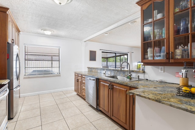 kitchen with dark stone countertops, glass insert cabinets, a sink, appliances with stainless steel finishes, and a textured ceiling