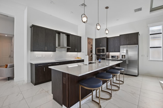 kitchen with pendant lighting, wall chimney range hood, sink, stainless steel appliances, and a center island with sink