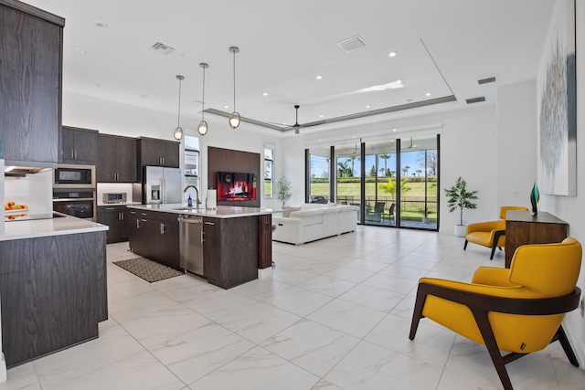 kitchen with a raised ceiling, appliances with stainless steel finishes, a center island with sink, and decorative light fixtures