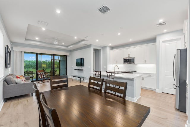 dining space with a raised ceiling, ceiling fan, sink, and light wood-type flooring