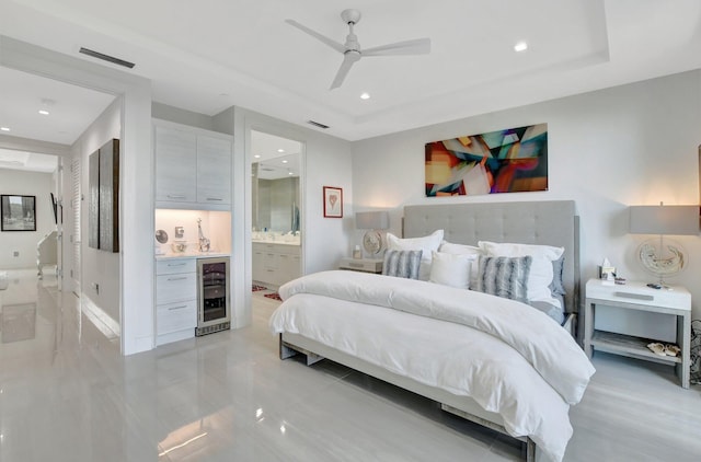 bedroom featuring wine cooler, ceiling fan, a tray ceiling, and connected bathroom