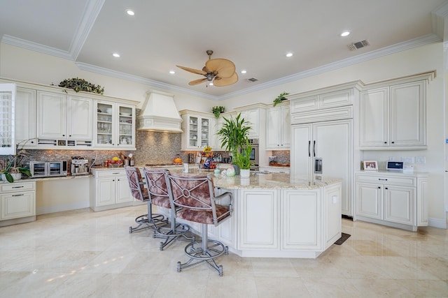 kitchen featuring tasteful backsplash, an island with sink, a kitchen breakfast bar, light stone counters, and custom range hood