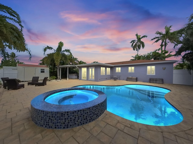 pool at dusk featuring an in ground hot tub, a storage unit, and a patio