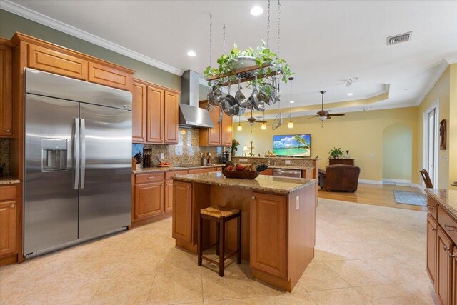 kitchen with decorative light fixtures, built in refrigerator, light stone counters, kitchen peninsula, and wall chimney range hood