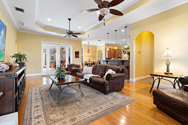 living room with a tray ceiling, light wood-style flooring, arched walkways, and ornamental molding