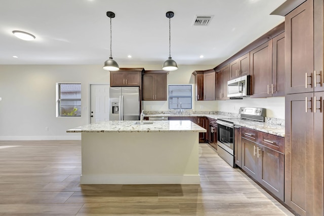 kitchen with dark brown cabinets, hanging light fixtures, an island with sink, stainless steel appliances, and light stone countertops