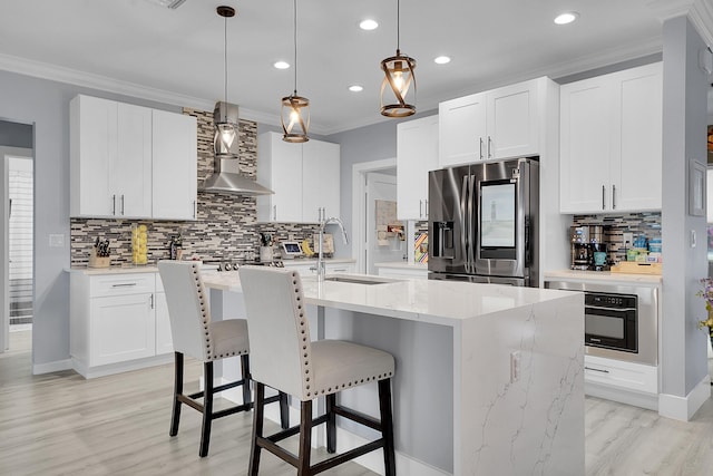 kitchen with white cabinetry, decorative light fixtures, a center island with sink, stainless steel fridge, and wall chimney range hood