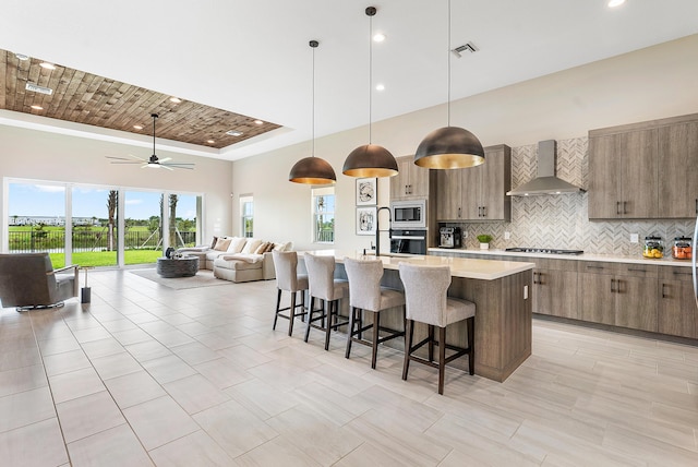 kitchen featuring wall chimney exhaust hood, decorative light fixtures, appliances with stainless steel finishes, an island with sink, and backsplash