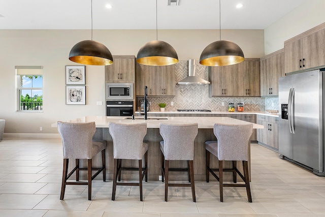 kitchen featuring wall chimney range hood, appliances with stainless steel finishes, backsplash, a center island with sink, and decorative light fixtures