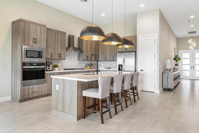 kitchen with sink, appliances with stainless steel finishes, a center island with sink, decorative light fixtures, and wall chimney exhaust hood