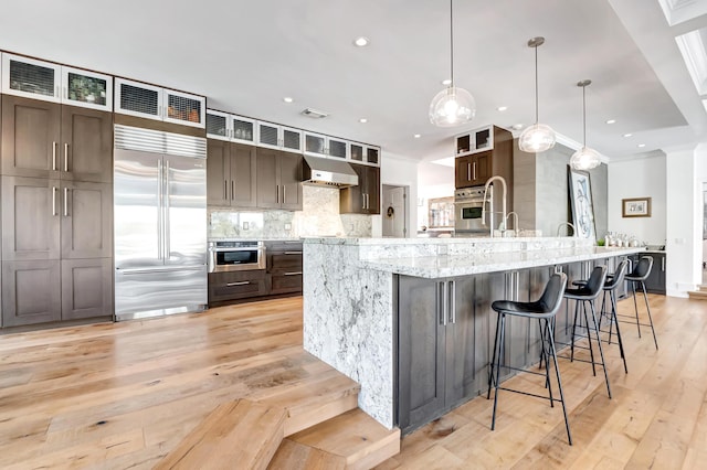 kitchen with decorative light fixtures, range hood, built in refrigerator, dark brown cabinets, and a spacious island