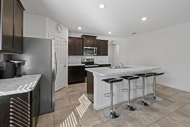 kitchen featuring arched walkways, appliances with stainless steel finishes, a sink, an island with sink, and dark brown cabinets