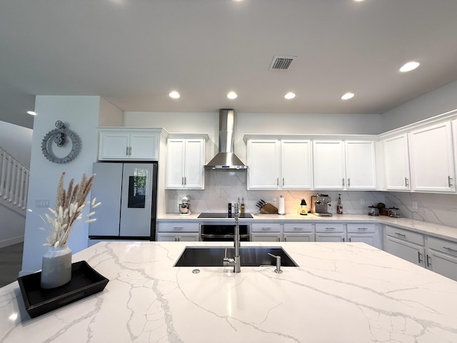 kitchen with stainless steel refrigerator, white cabinetry, light stone counters, and wall chimney range hood