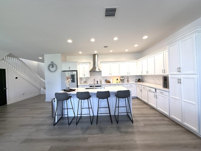 kitchen featuring stainless steel refrigerator, an island with sink, wall chimney range hood, decorative backsplash, and white cabinets