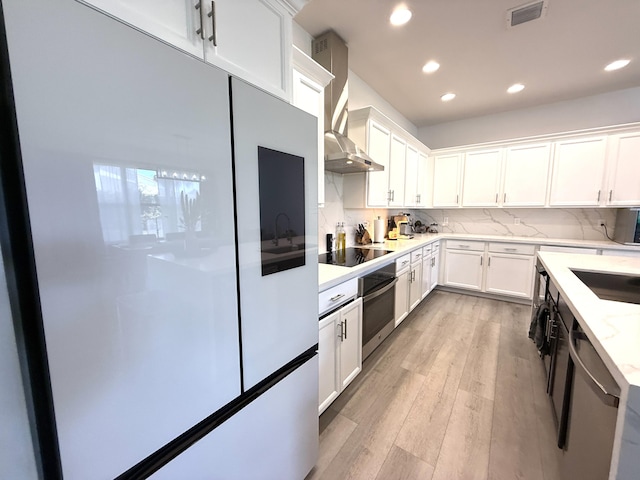 kitchen with wall chimney exhaust hood, white cabinetry, appliances with stainless steel finishes, light hardwood / wood-style floors, and decorative backsplash