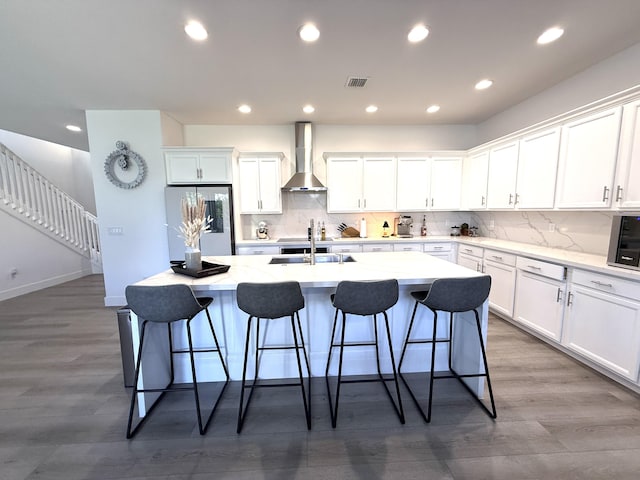 kitchen with white cabinetry, wall chimney range hood, fridge, and a center island with sink