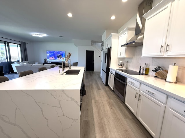 kitchen with an island with sink, sink, white cabinets, stainless steel appliances, and wall chimney range hood