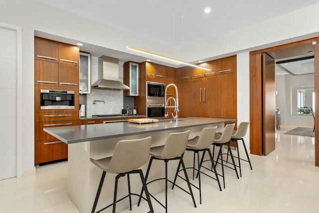 kitchen with stainless steel appliances, backsplash, brown cabinetry, modern cabinets, and wall chimney exhaust hood