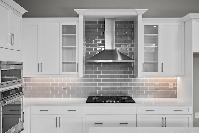 kitchen with appliances with stainless steel finishes, wall chimney range hood, white cabinets, and backsplash