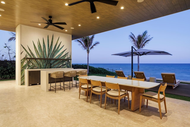 patio terrace at dusk with ceiling fan, a water view, and a bar