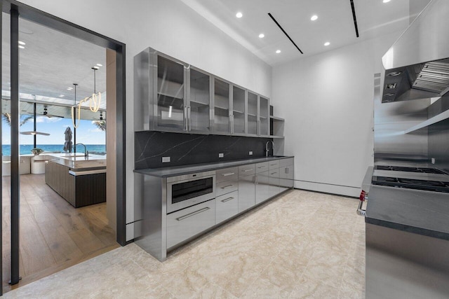 kitchen with a water view, tasteful backsplash, an island with sink, stainless steel microwave, and ventilation hood