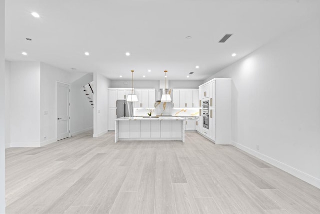 kitchen featuring pendant lighting, wall chimney range hood, a kitchen island with sink, white cabinets, and light wood-type flooring