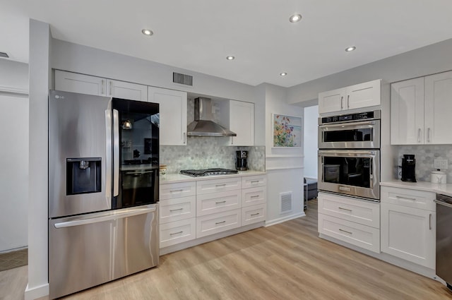 kitchen with wall chimney range hood, backsplash, stainless steel appliances, light hardwood / wood-style floors, and white cabinets