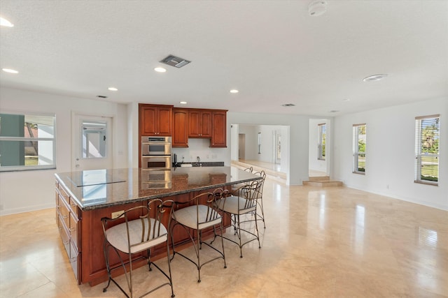 kitchen with a breakfast bar, cooktop, stainless steel double oven, dark stone counters, and a large island