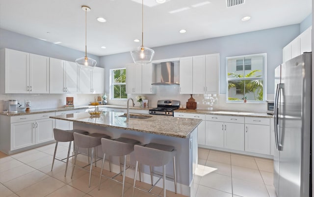 kitchen featuring sink, wall chimney range hood, appliances with stainless steel finishes, light stone countertops, and an island with sink