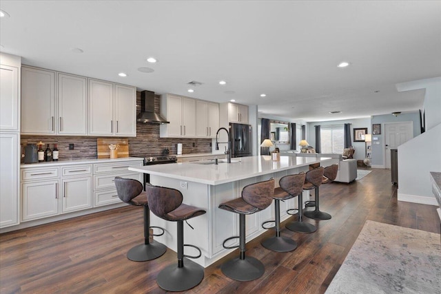 kitchen with dark wood-type flooring, sink, an island with sink, decorative backsplash, and wall chimney range hood