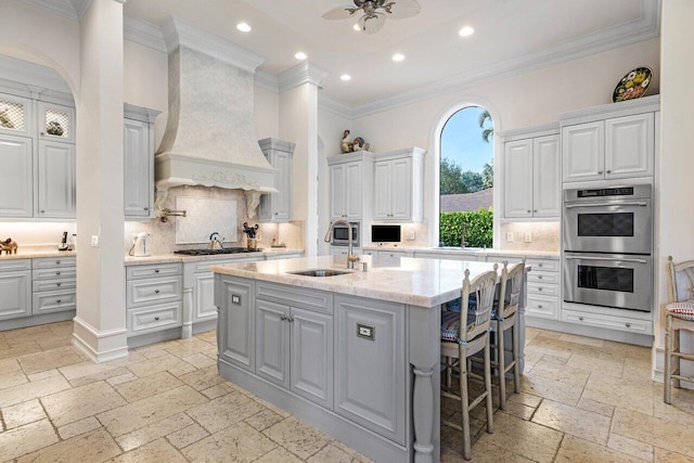 kitchen featuring stone tile floors, custom range hood, double oven, a sink, and a kitchen island with sink