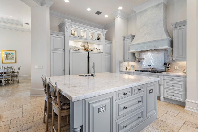 kitchen featuring crown molding, baseboards, stone tile flooring, a center island with sink, and custom range hood