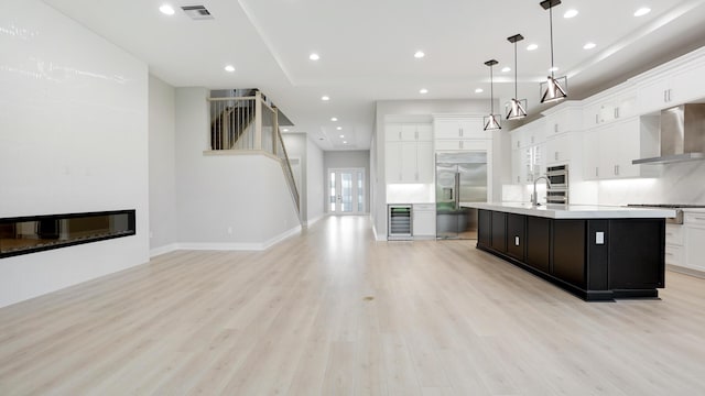 kitchen with pendant lighting, wall chimney range hood, appliances with stainless steel finishes, white cabinetry, and an island with sink