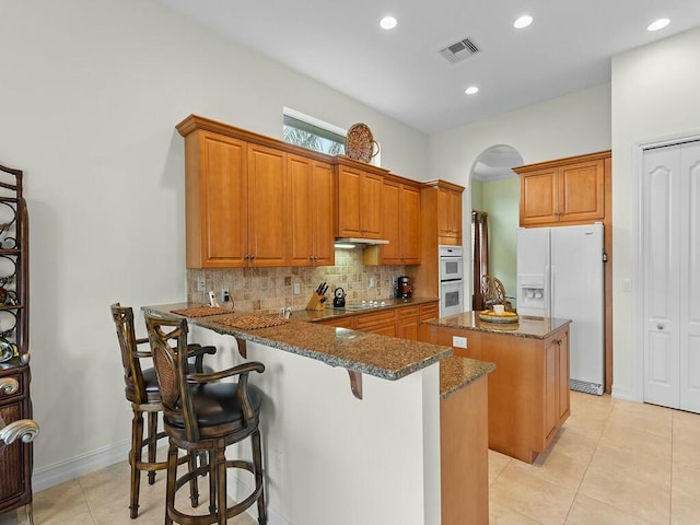 kitchen featuring a breakfast bar, a center island, kitchen peninsula, white appliances, and dark stone counters