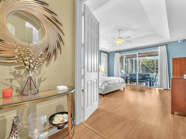 bedroom featuring light hardwood / wood-style flooring, access to outside, a textured ceiling, a raised ceiling, and ceiling fan