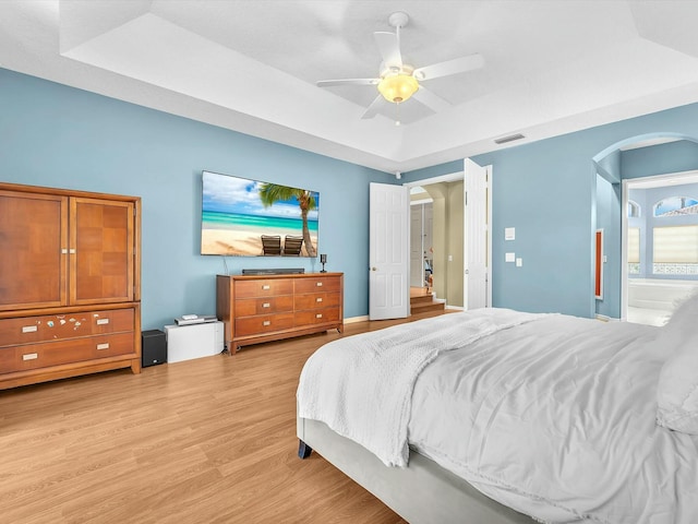 bedroom featuring ceiling fan, connected bathroom, light hardwood / wood-style floors, and a tray ceiling