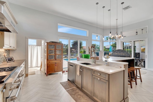 kitchen with pendant lighting, wall chimney range hood, sink, stainless steel appliances, and a center island with sink