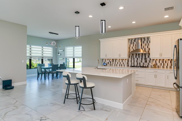 kitchen featuring pendant lighting, white cabinets, backsplash, a kitchen island with sink, and wall chimney exhaust hood