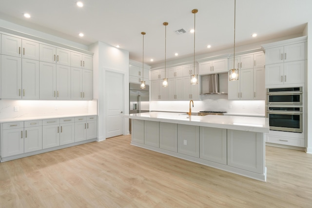 kitchen featuring pendant lighting, appliances with stainless steel finishes, light hardwood / wood-style floors, a center island with sink, and wall chimney exhaust hood