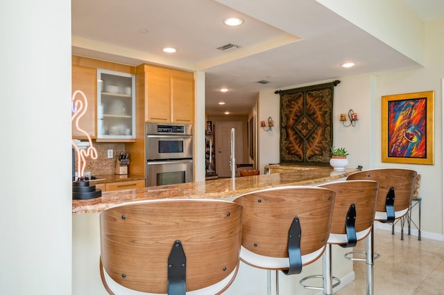 kitchen with light brown cabinetry, a kitchen breakfast bar, decorative backsplash, light stone countertops, and stainless steel double oven