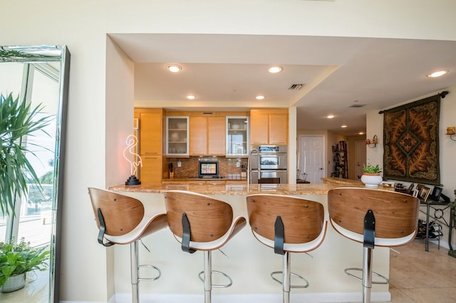 kitchen with a breakfast bar, light brown cabinetry, kitchen peninsula, stainless steel double oven, and decorative backsplash