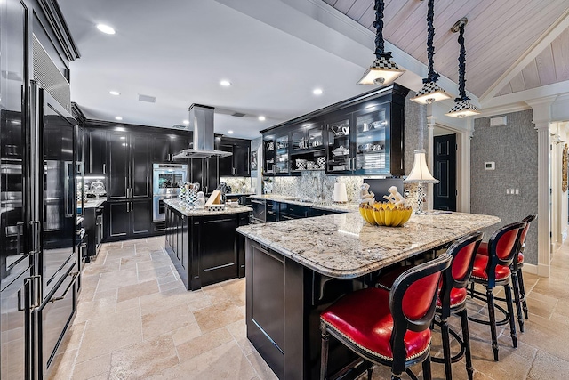 kitchen with a center island, light stone counters, island range hood, black built in refrigerator, and decorative light fixtures