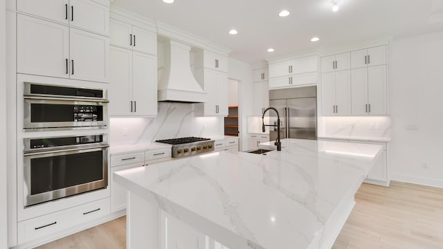 kitchen with a center island with sink, light stone countertops, custom range hood, and appliances with stainless steel finishes